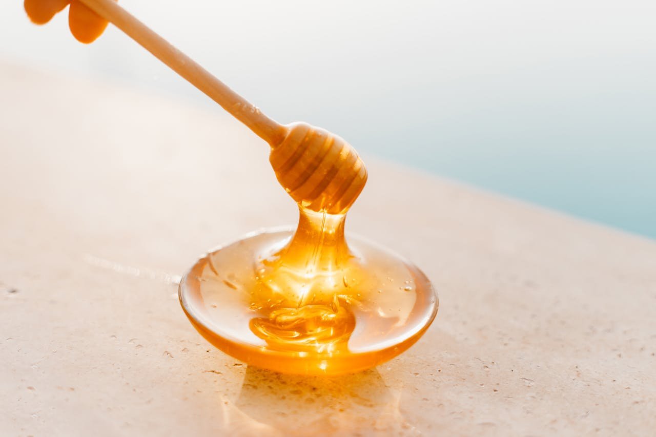 about-us-02 Golden honey flowing from a wooden dipper onto a glass plate, captured in warm daylight.